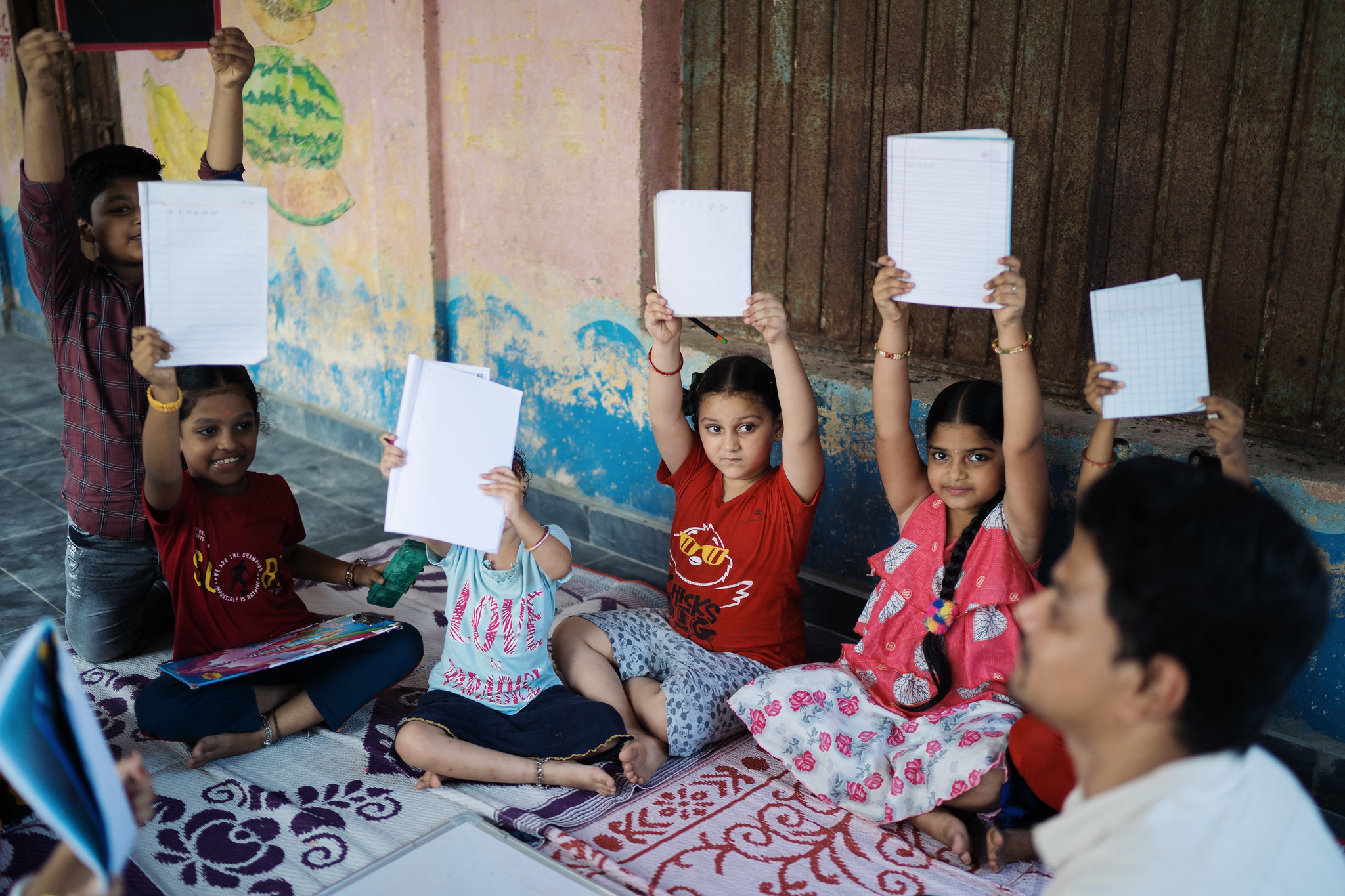 Children sitting in a circle holding school notebooks in the air.